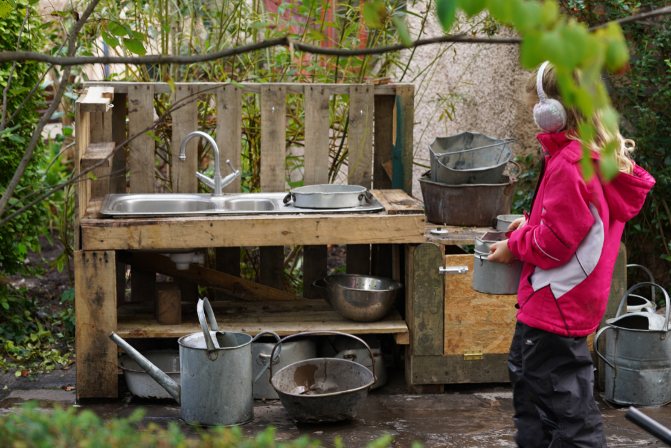 A young girl playig at a mud kitchen outdoors, in kindergarten nursery setting, carrying water in pot. Alternative envirnment for 4yo instead of P1 classroom, excercising statutory right to defer entry to P1